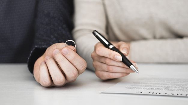 One hand holding a ring, another hand holding a pen over a divorce agreement.